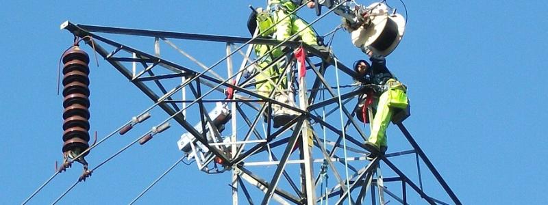 Höghöjdsarbete pågår: Nya datakablar installeras på master. Högt uppe på en elektricitetsmast arbetar kablagearbetare med att lägga till en datakabel. De använder en specialmaskin som rullar upp kabeln på en trumma medan den färdas längs stödkabeln som sträcker sig mellan tornen. Maskinen, som drivs av egen motor, rör sig självständigt från ett torn till ett annat. När den når ett torn monteras den ner, flyttas över till andra sidan och fortsätter sedan sin färd. På bilden har drivmotorn redan flyttats över, men kabeltrumman väntar fortfarande på att hissas över tornet. Foto: Wikipedia Common, kredit: Sandpiper